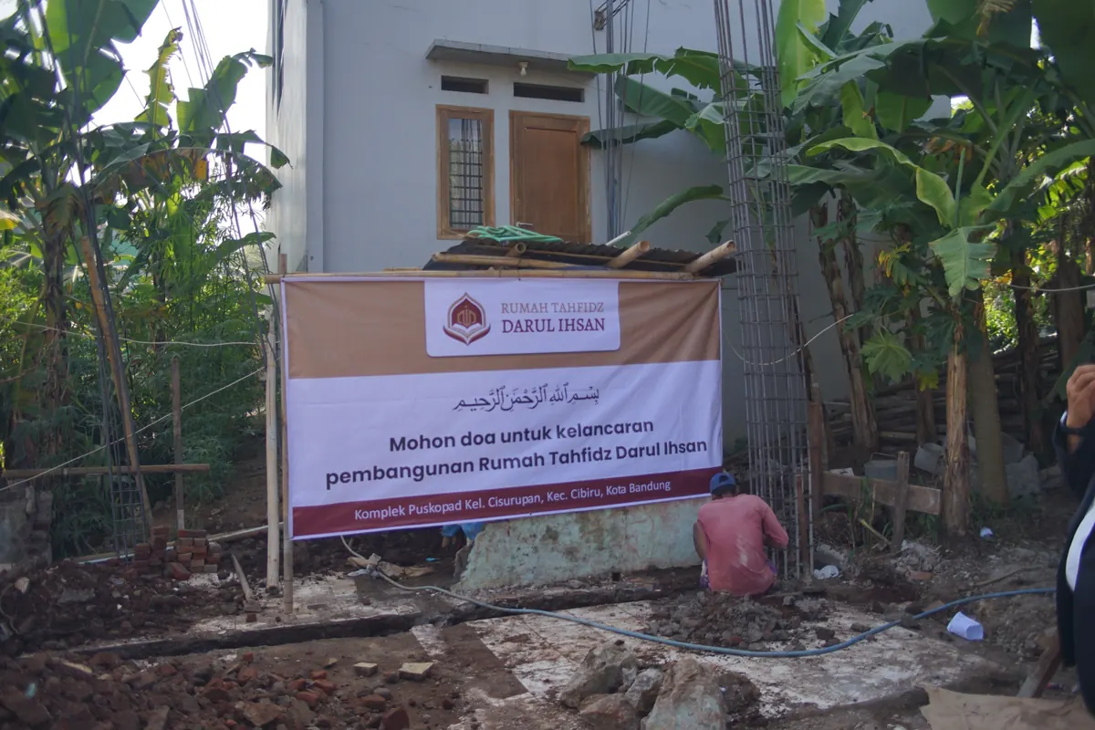 Lokasi pembangunan dengan tumpukan bata merah, rangka besi tulangan kolom, dan spanduk pembangunan Rumah Tahfidz