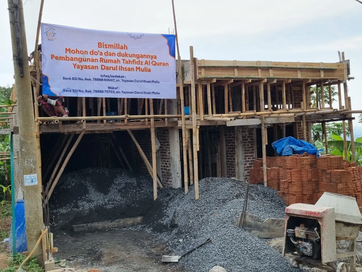 Areal kerja pembangunan dengan perancah bambu, tumpukan kerikil, molen, dan spanduk di latar belakang