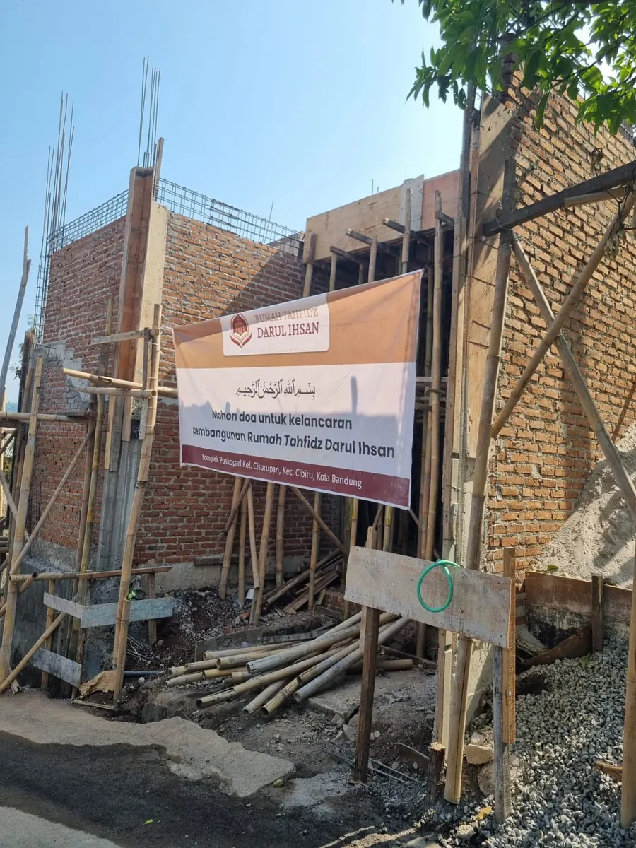 Struktur dinding bata merah dengan penyangga kayu dan spanduk pembangunan terpasang di lokasi